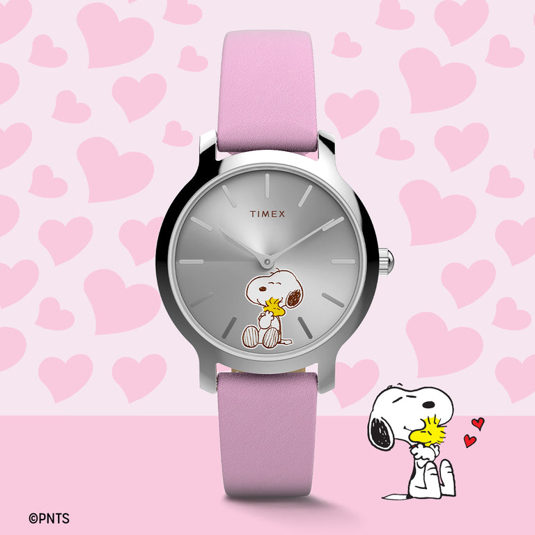 Timex Peanuts® watch with a silver-tone case, light pink leather strap, and a silver dial featuring Snoopy hugging Woodstock, displayed against a pink background with heart graphics.