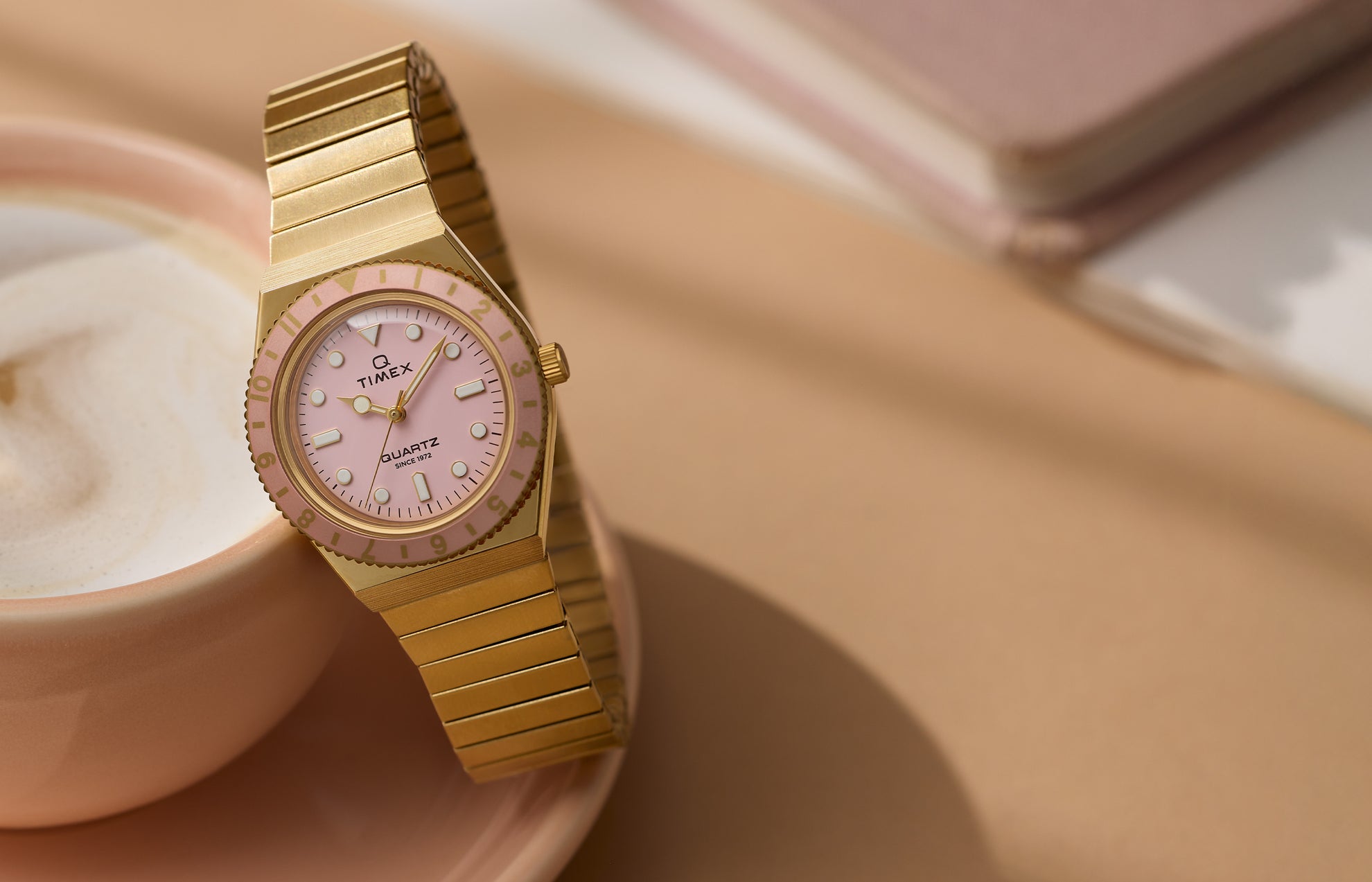 Q Timex® Mini gold-tone watch with expansion bracelet, pale pink dial, and matching pink rotating bezel, styled beside a cup of coffee.