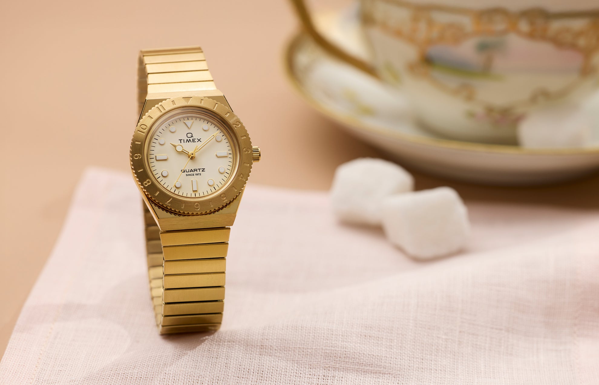 Q Timex® Mini gold-tone watch featuring a cream dial, rotating bezel, and gold expansion bracelet, displayed on a soft neutral tabletop.