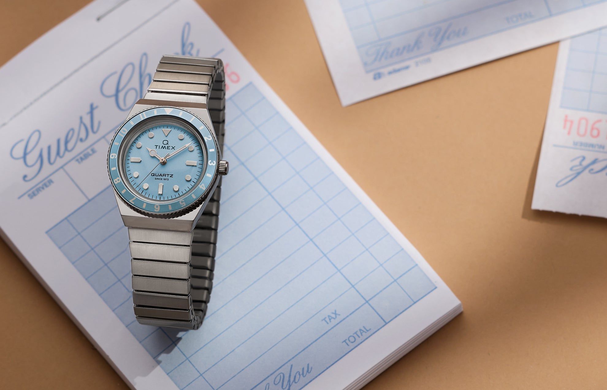 Q Timex® Mini silver-tone watch with light blue dial, rotating bezel, and stainless steel expansion bracelet, styled on a guest check pad.