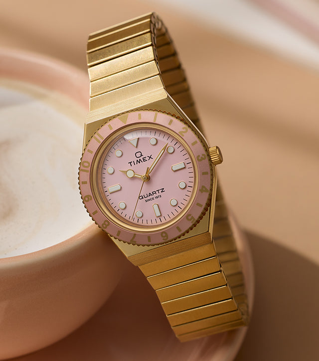 Q Timex® Mini gold-tone watch with expansion bracelet, pale pink dial, and matching pink rotating bezel, styled beside a cup of coffee.