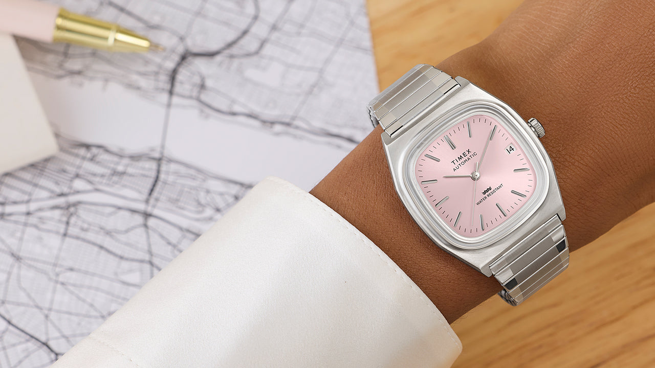 Timex® Automatic 1983 E Line watch with light pink dial and stainless steel expansion bracelet worn on wrist near a drafting sketch.