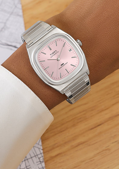 Timex® Automatic 1983 E Line watch with light pink dial and stainless steel expansion bracelet worn on wrist near a drafting sketch.
