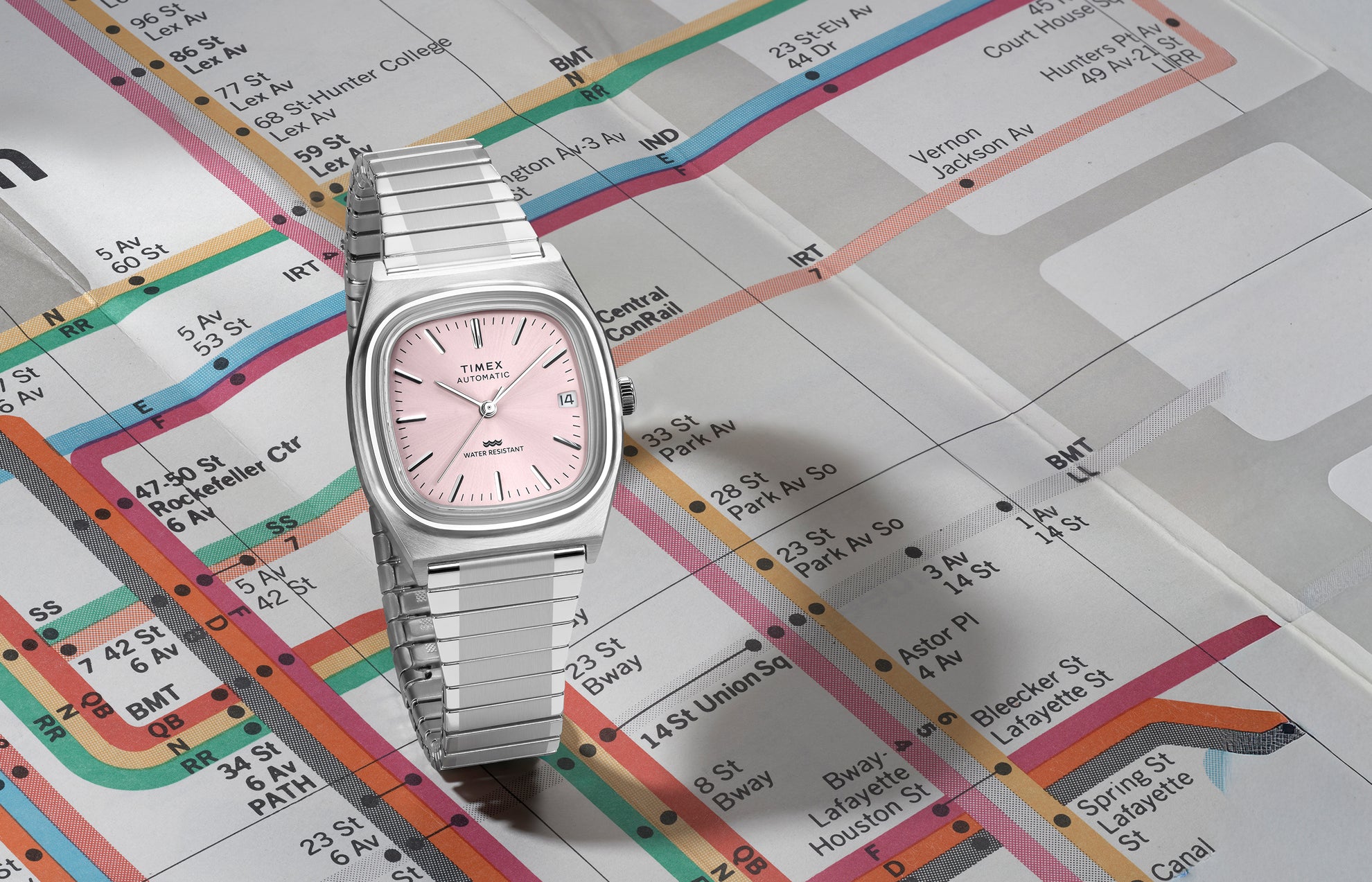 Timex® Automatic 1983 E Line stainless steel watch with light pink sunray dial displayed at an angle on a vintage subway map.