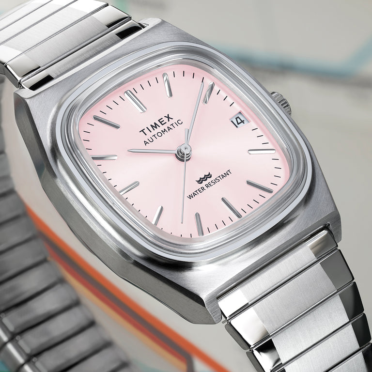 Close-up of the Timex® Automatic 1983 E Line watch showing a light pink sunray dial, applied silver hour markers, date window, and brushed stainless steel case.