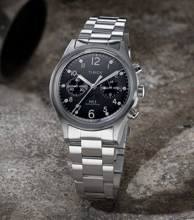 Timex MK1 Chronograph with stainless steel bracelet and black dial, shown standing upright on a textured stone surface.