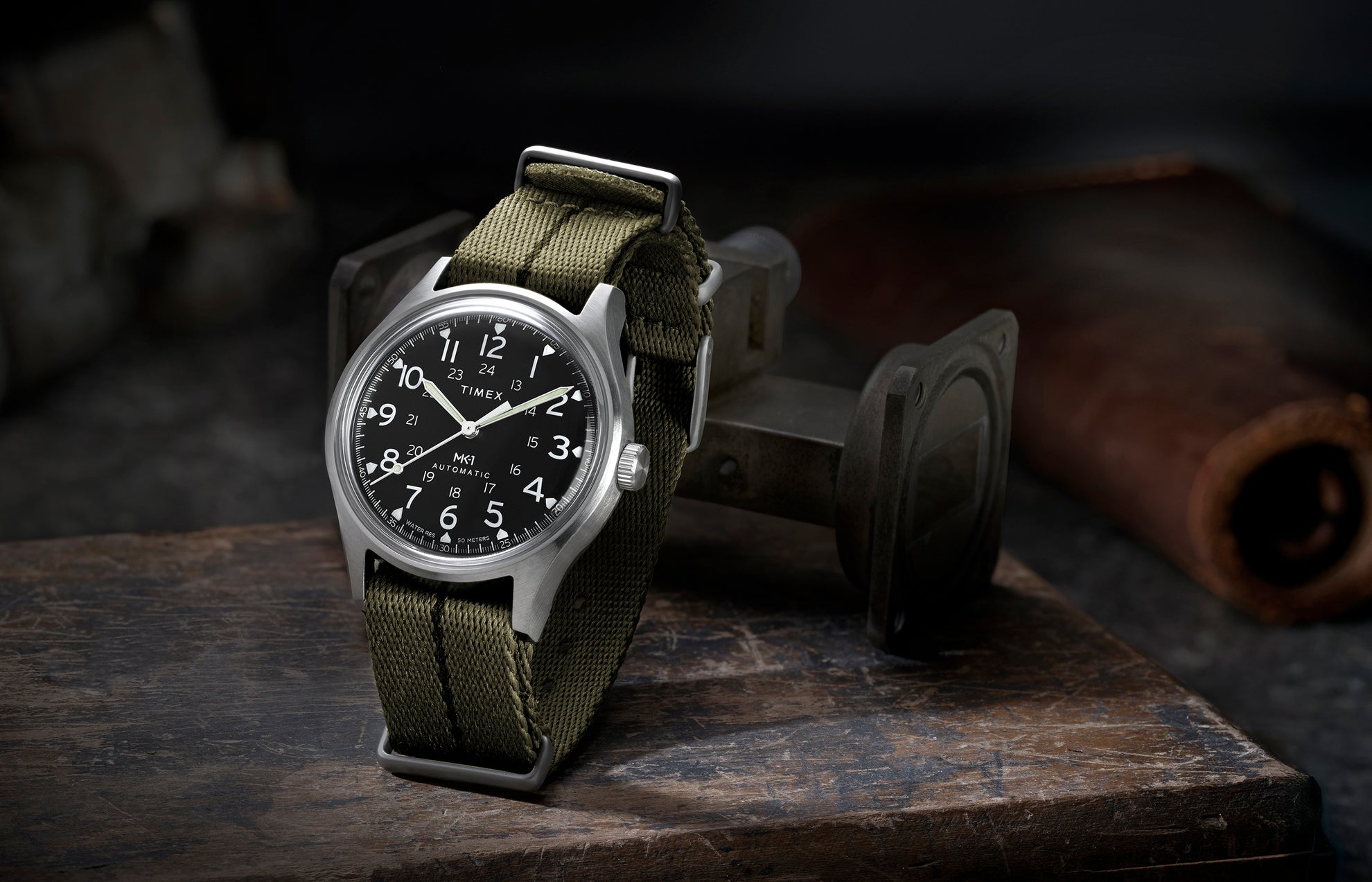 Timex MK1 Automatic with green fabric strap and black dial, displayed upright on a rugged industrial surface.