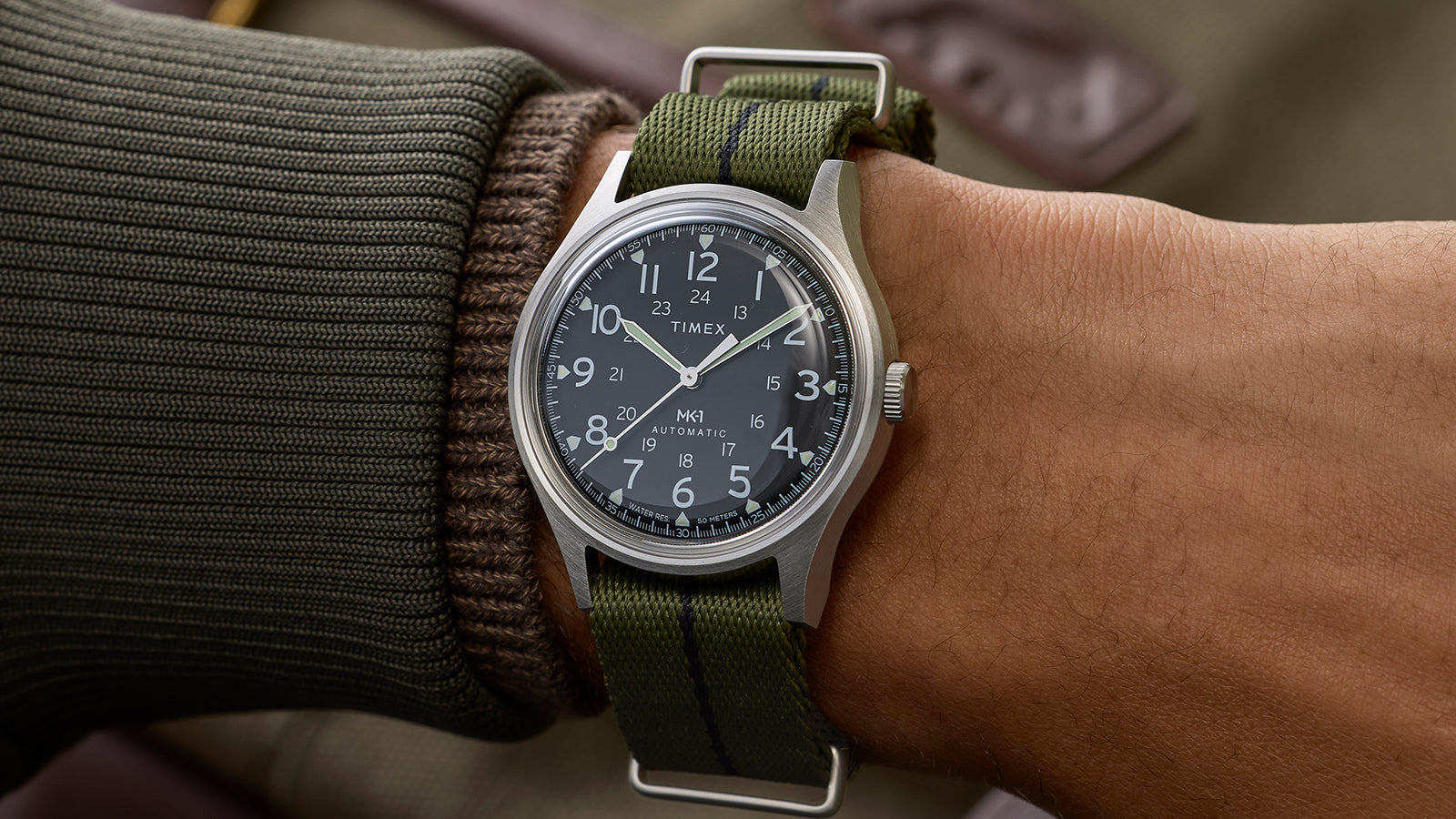 Timex MK1 Automatic worn on wrist with green fabric strap, stainless steel case, and black field-style dial.
