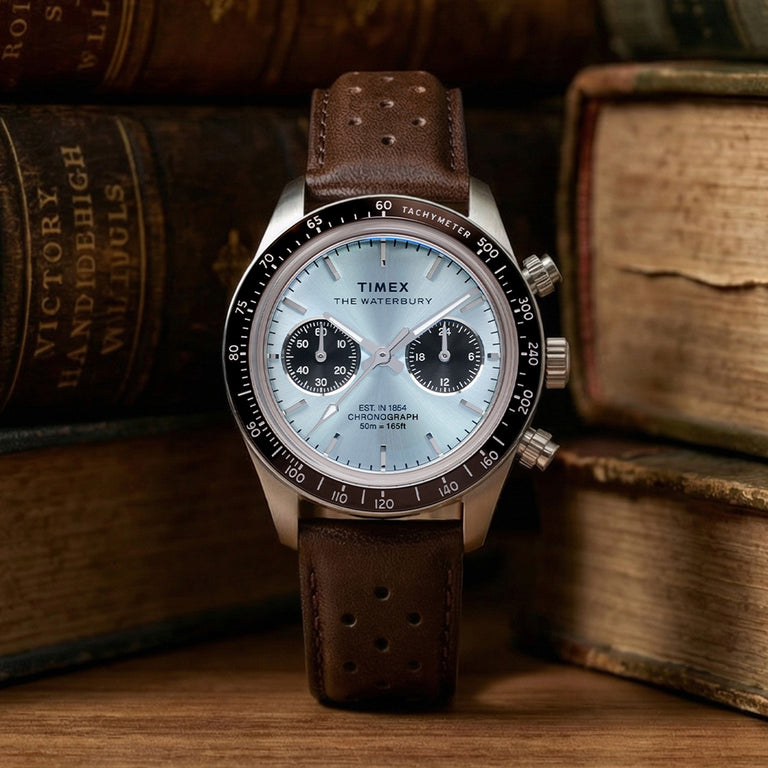 Timex Waterbury Heritage Chronograph with brown leather strap and light blue dial, shown front-facing against a vintage backdrop.