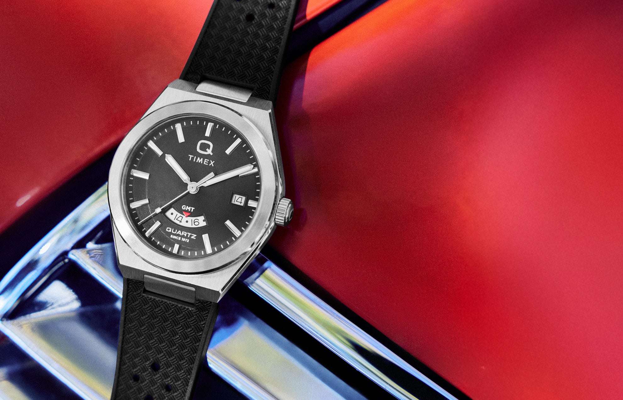 Q Timex Continental GMT watch with a black dial and silver stainless steel case, featuring a date window and GMT subdial, shown on a textured black rubber strap against a bold red and metallic background.