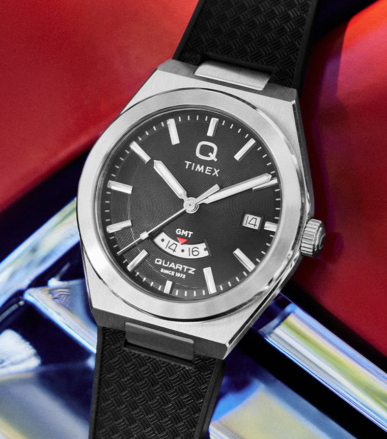Q Timex Continental GMT watch with a black dial and silver stainless steel case, featuring a date window and GMT subdial, shown on a textured black rubber strap against a bold red and metallic background.