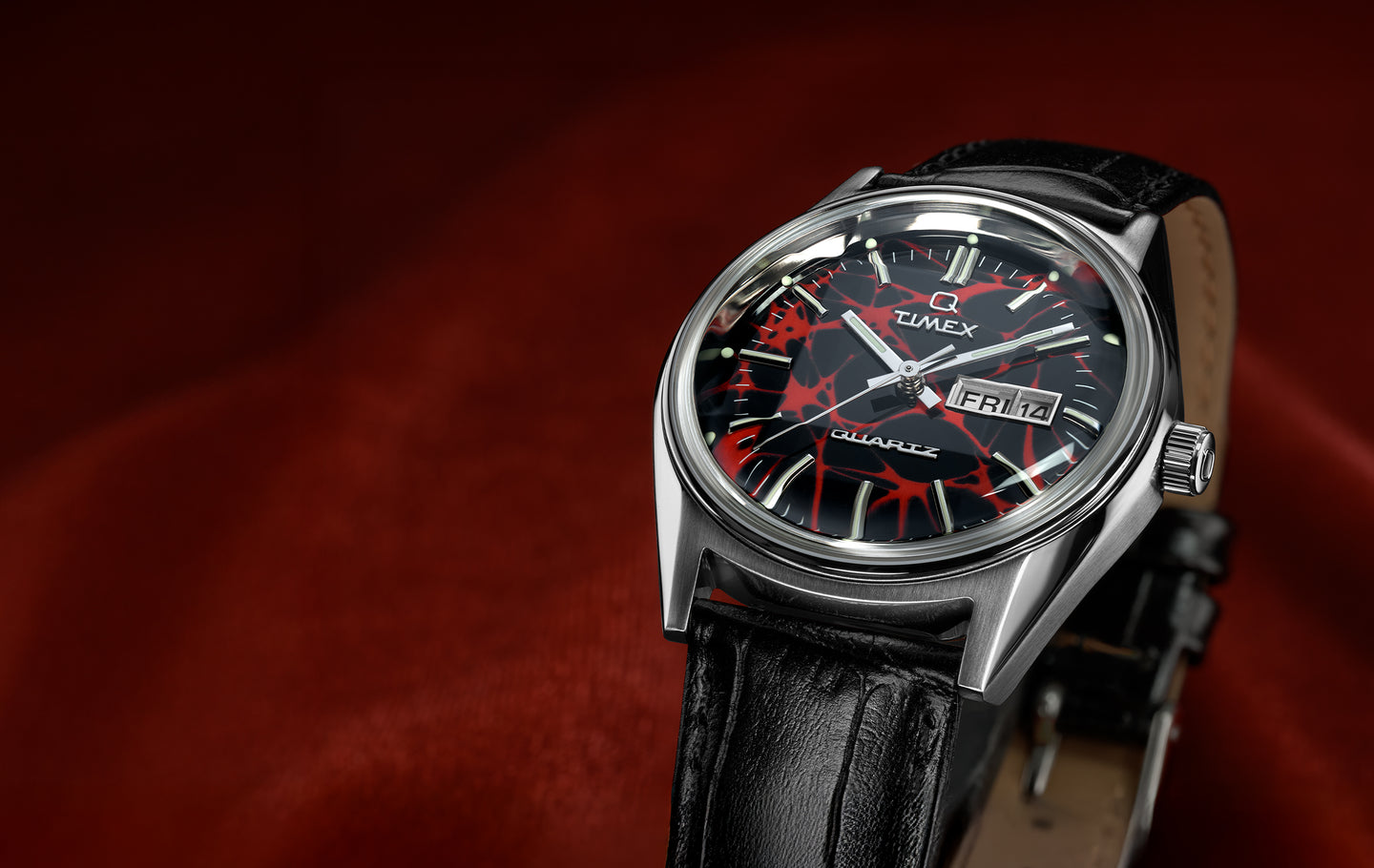 Q Timex Red Vega 1978 Reissue watch with a stainless steel case, black leather strap, and a black dial featuring a bold red web-like pattern, luminous hour markers, silver hands, and a day-date window, shown against a deep red background.