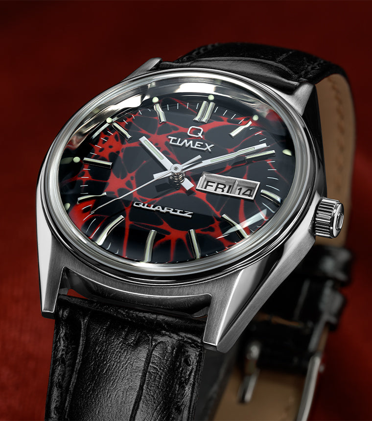 Q Timex Red Vega 1978 Reissue watch with a stainless steel case, black leather strap, and a black dial featuring a bold red web-like pattern, luminous hour markers, silver hands, and a day-date window, shown against a deep red background.