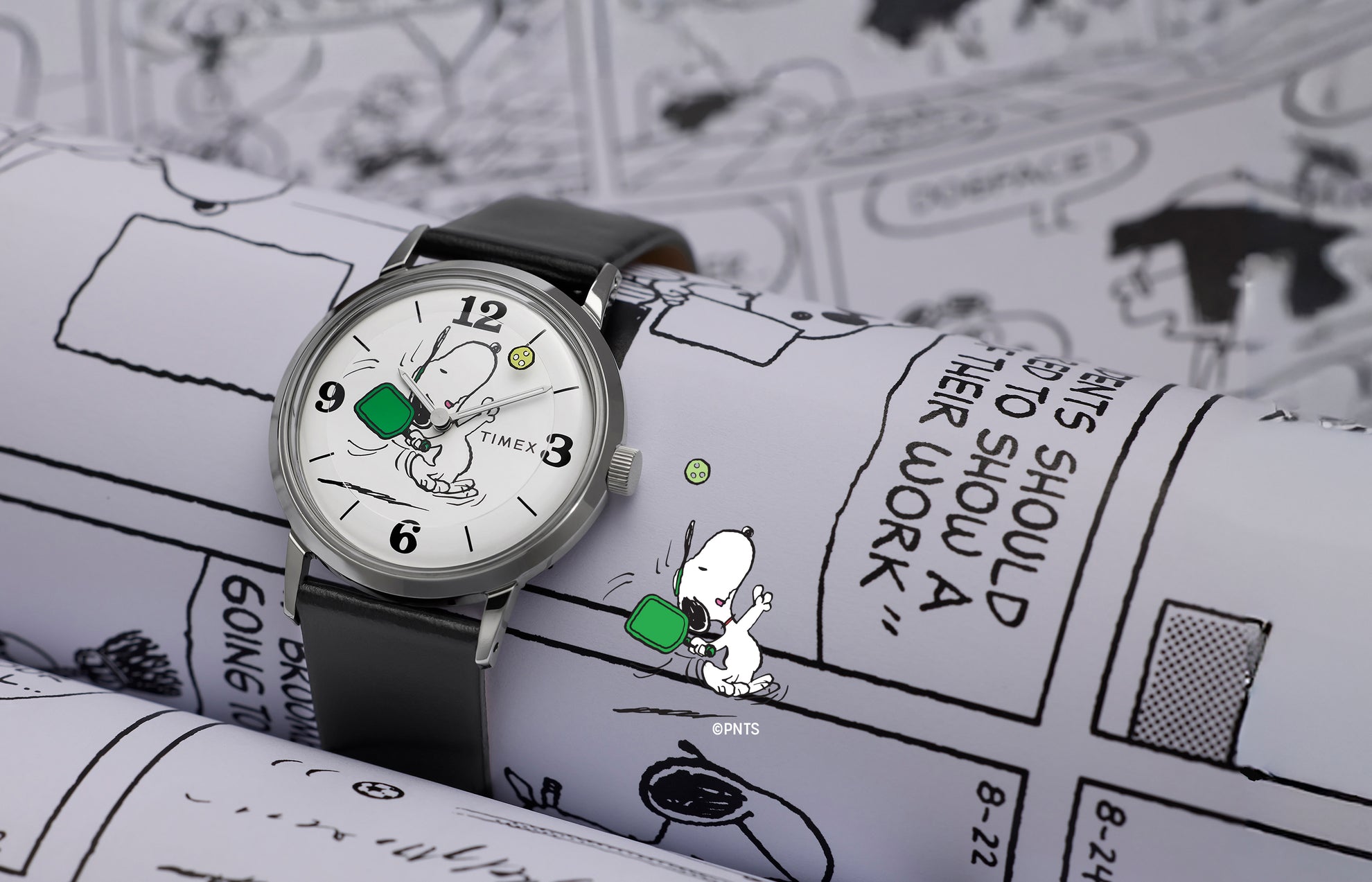 Peanuts® x Timex Marlin® Pickleball watch with black leather strap and white dial featuring Snoopy playing pickleball, displayed on Peanuts comic strip background.