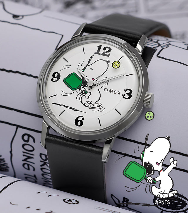 Peanuts® x Timex Marlin® Pickleball watch with black leather strap and white dial featuring Snoopy playing pickleball, displayed on Peanuts comic strip background.