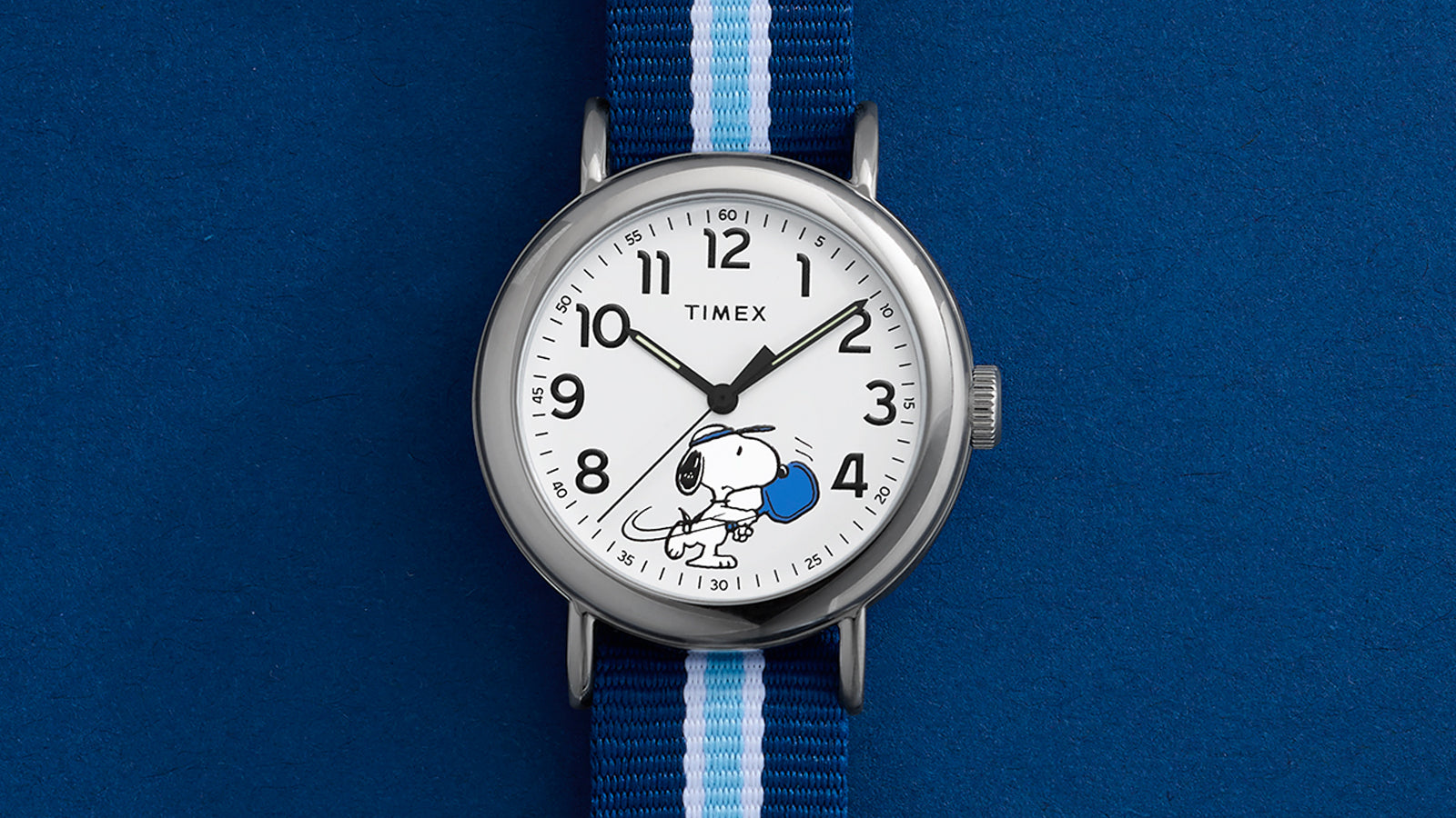 Close-up of Peanuts® x Timex Weekender® Pickleball dial showing Snoopy swinging a blue pickleball paddle, black Arabic numerals, and minute track.