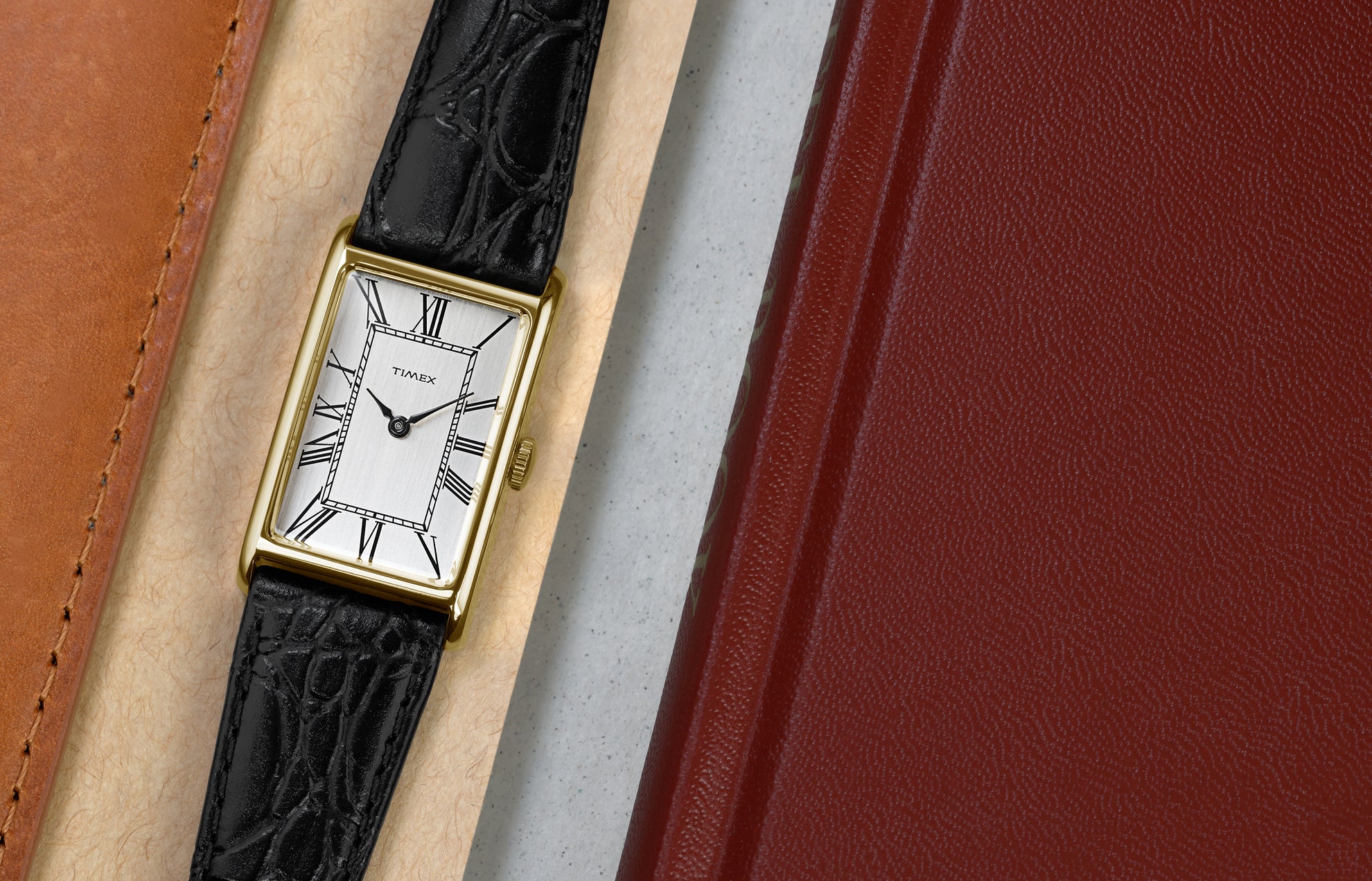 Timex 1976 Lexington Reissue watch laid flat beside leather accessories, highlighting the slim gold-tone case and classic Roman numeral dial.