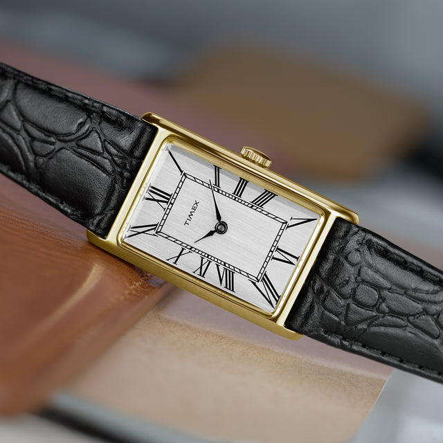 Close-up of the Timex 1976 Lexington Reissue featuring a gold-tone rectangular case, brushed silver dial, black Roman numerals, and black leather strap resting on a leather surface.