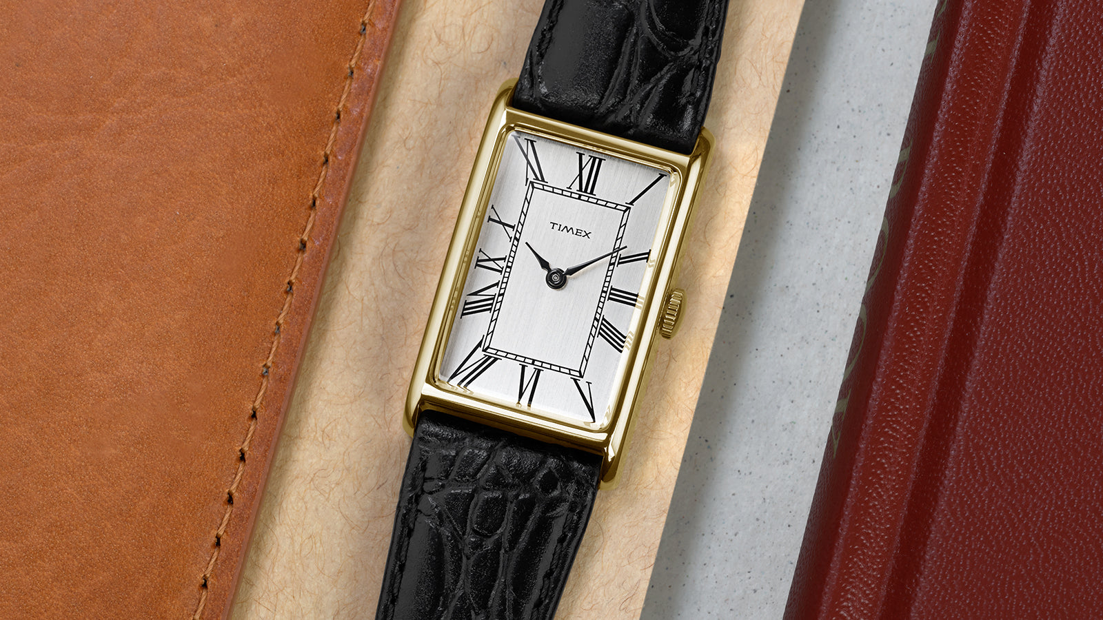 Timex 1976 Lexington Reissue watch laid flat beside leather accessories, highlighting the slim gold-tone case and classic Roman numeral dial.