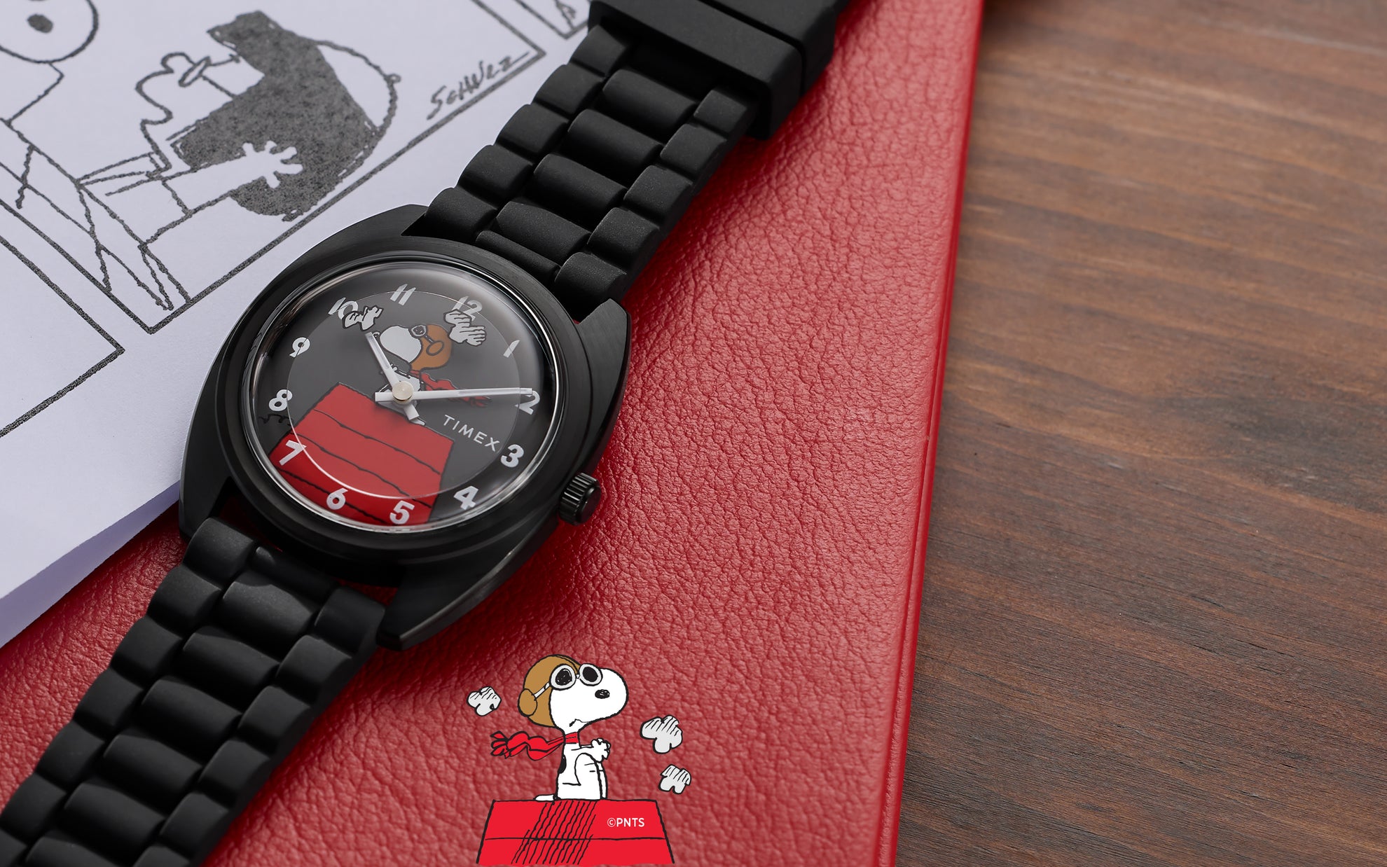 Peanuts® x Timex 1979 Reissue Inspired watch with black bracelet and gray dial showcasing Snoopy Flying Ace artwork, placed on a red notebook beside Peanuts comic strip pages.