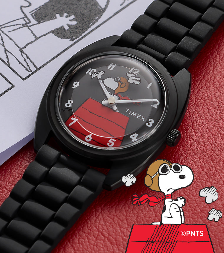 Peanuts® x Timex 1979 Reissue watch with a black case and bracelet, featuring Snoopy flying his red doghouse on the dial, placed on a red surface beside a Peanuts comic strip page.