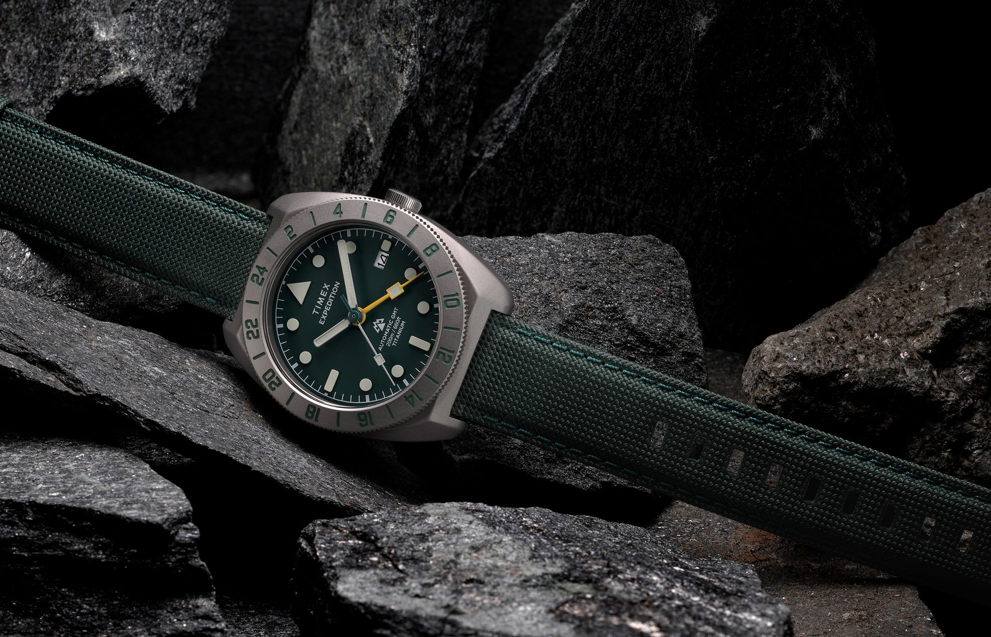 Timex Expedition Pioneer Titanium Automatic GMT watch with green dial and fabric strap resting on dark rocks.