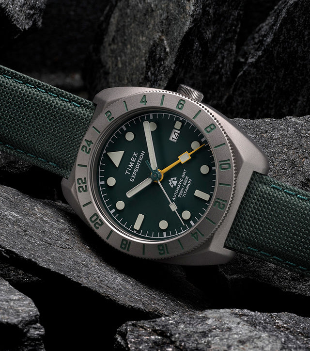 Timex Expedition Pioneer Titanium Automatic GMT watch with green dial and fabric strap resting on dark rocks.