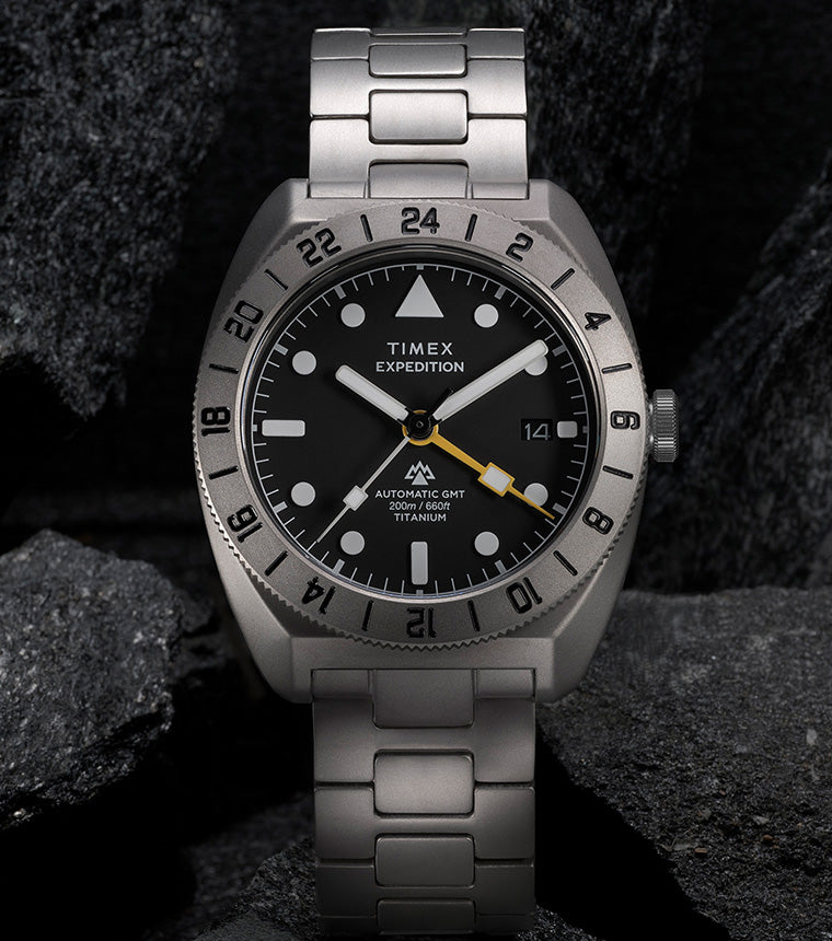 Expedition® Pioneer Titanium Automatic GMT standing upright on rocks, showcasing the titanium bracelet, black dial, and engraved 24-hour bezel.