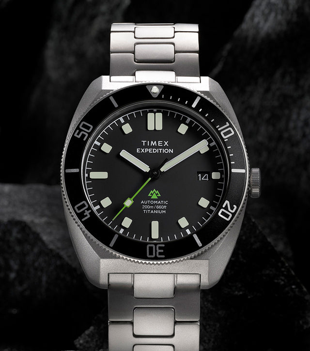 Expedition® Pioneer Titanium Automatic standing on rugged rocks, highlighting the titanium bracelet, black dial, rotating bezel, and date window.