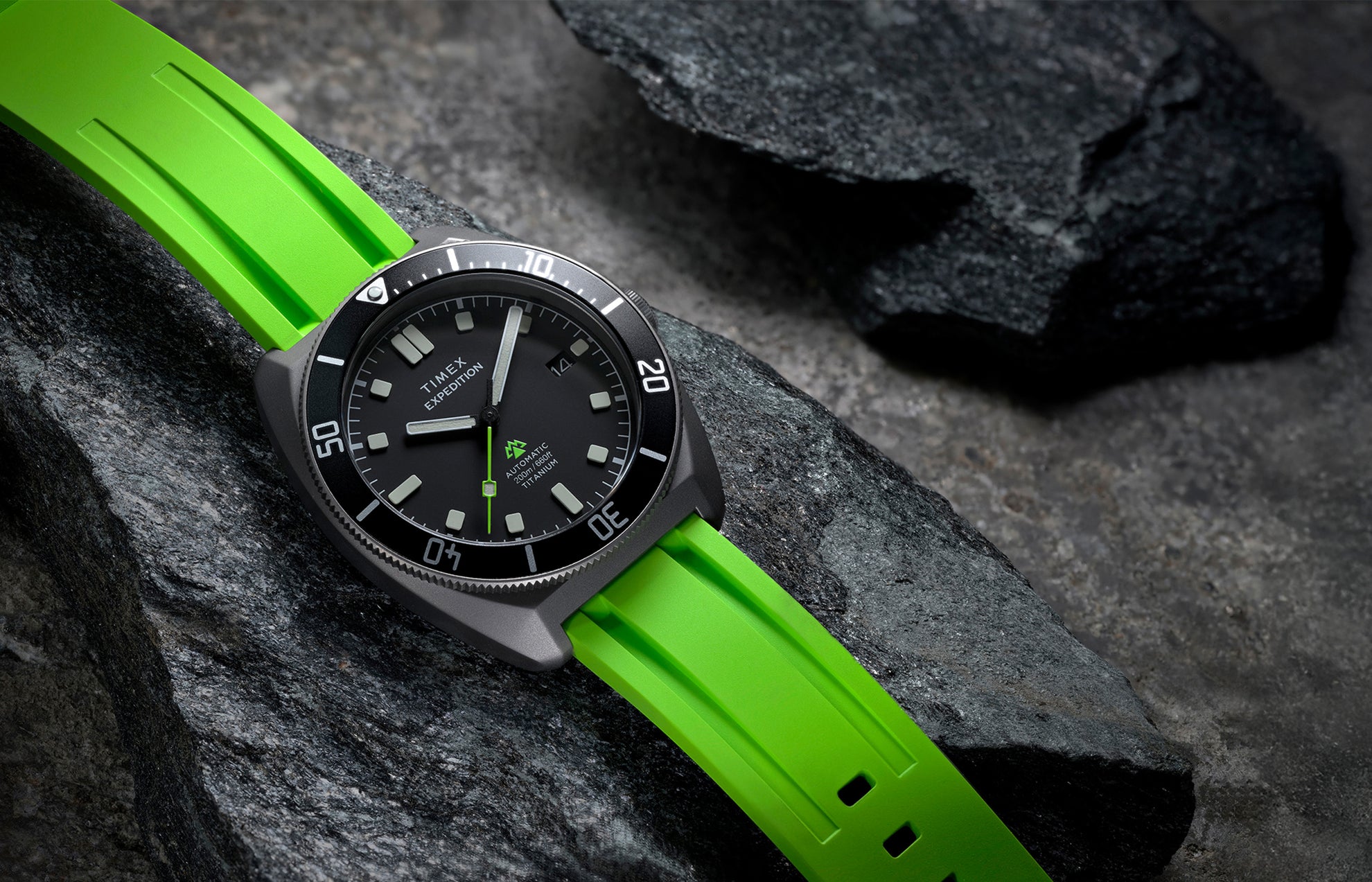Expedition® Pioneer Titanium Automatic resting on rugged rocks, highlighting the black dial, rotating bezel, and vivid green strap.
