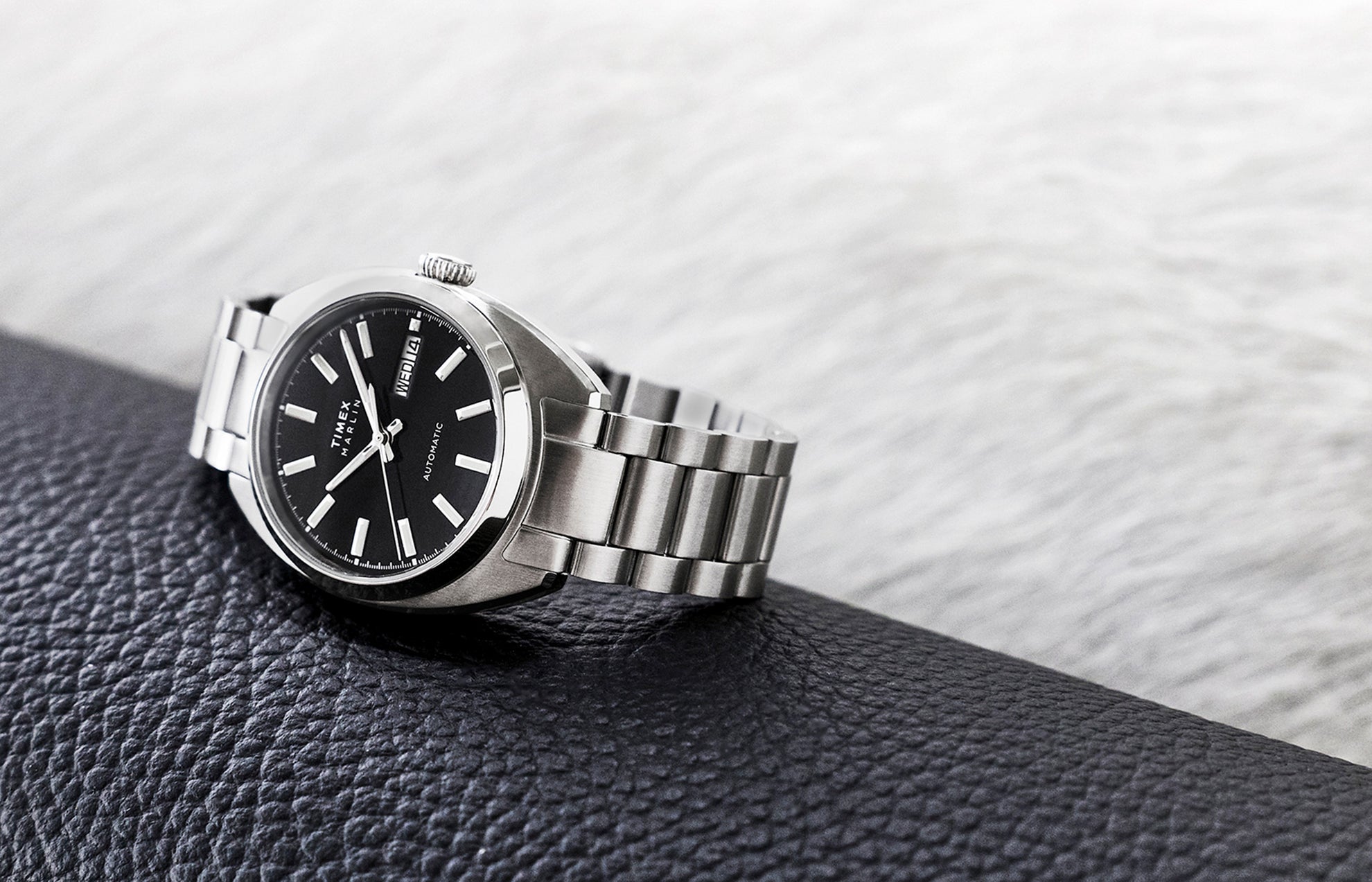 Timex Marlin Draper Automatic stainless steel watch resting on a textured surface, featuring a black dial and metal bracelet.