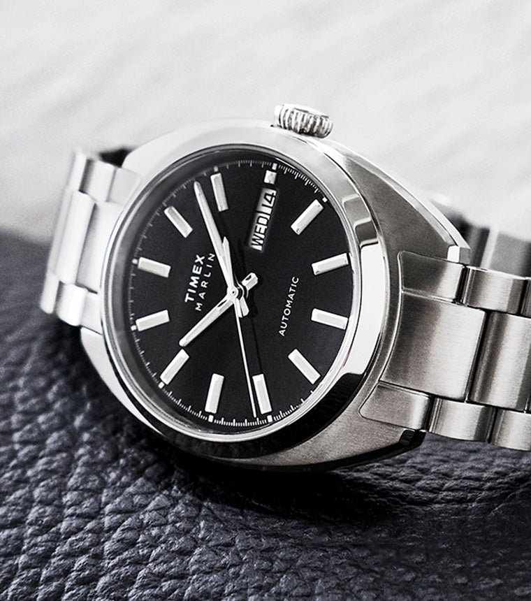 Timex Marlin Draper Automatic stainless steel watch resting on a textured surface, featuring a black dial and metal bracelet.