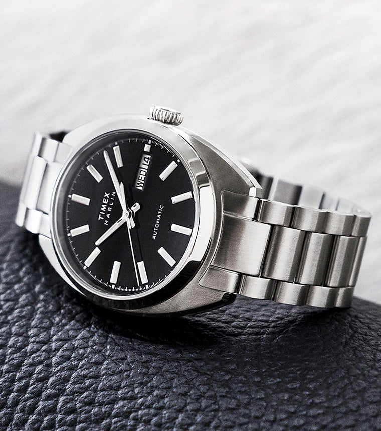 Timex Marlin® Draper Automatic stainless steel watch with black dial and silver bracelet resting on a textured black surface.