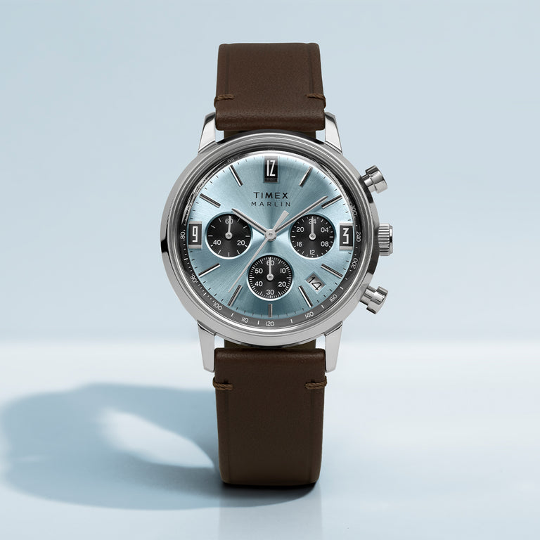 Timex Marlin Chronograph Tachymeter watch with blue dial, stainless steel case, and brown leather strap on light blue background.