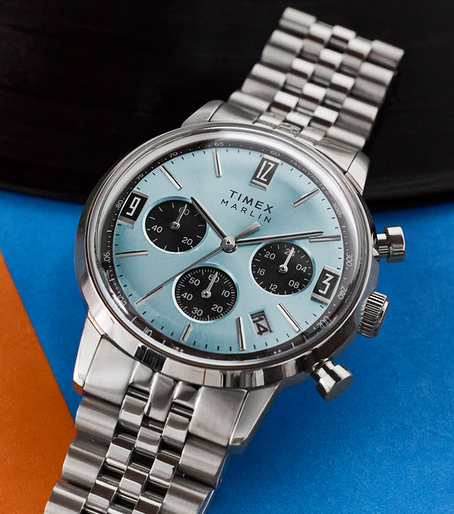 Timex Marlin Chronograph Tachymeter watch with a light blue dial, black chronograph subdials, and stainless steel bracelet, resting on a blue and orange graphic surface.