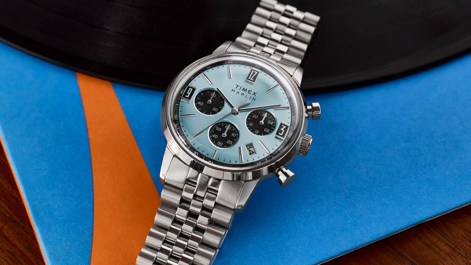 Side profile of the Timex Marlin Chronograph Tachymeter showing the stainless steel case, crown, pushers, and bracelet against a blue and orange background.