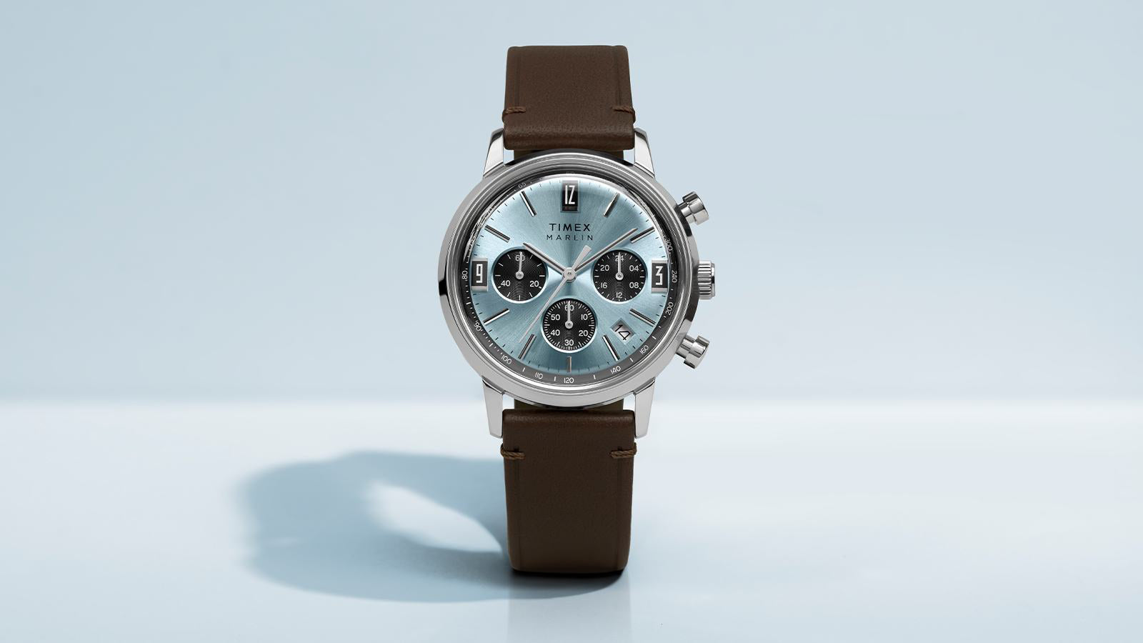 Timex Marlin Chronograph Tachymeter watch with a light blue dial, black subdials, stainless steel case, and brown leather strap, shown front-facing on a light background.