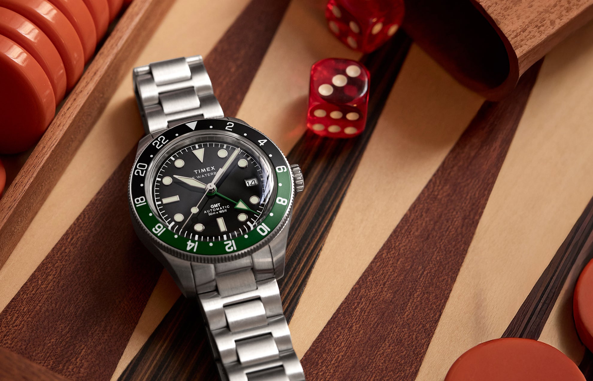 Timex Waterbury Heritage Automatic GMT watch with green and black bezel on a wooden game board with red dice.