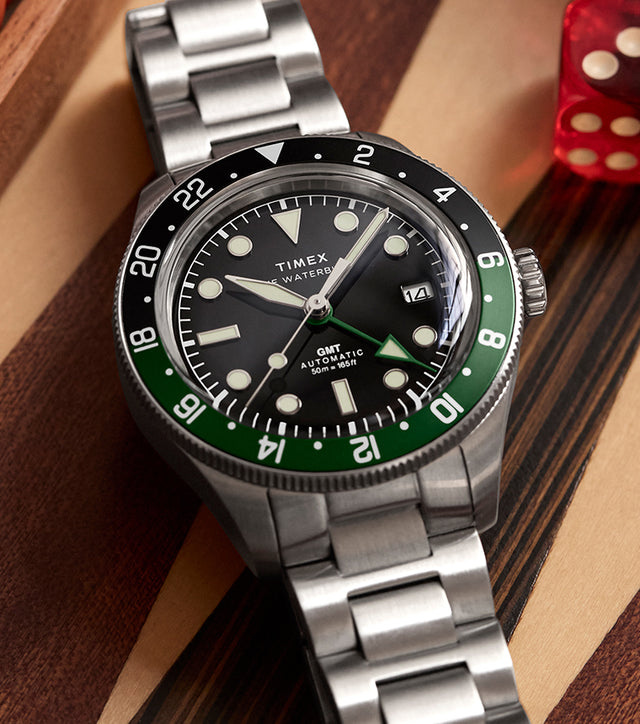 Timex Waterbury Heritage Automatic GMT watch with green and black bezel on a wooden game board with red dice.