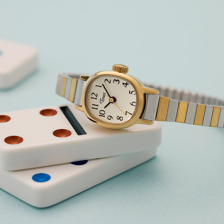 Timex Cavatina women's dress watch featuring a gold-tone case, white dial with full Arabic numerals, and a two-tone stainless-steel expansion bracelet, styled on white dominoes against a pastel blue backdrop.