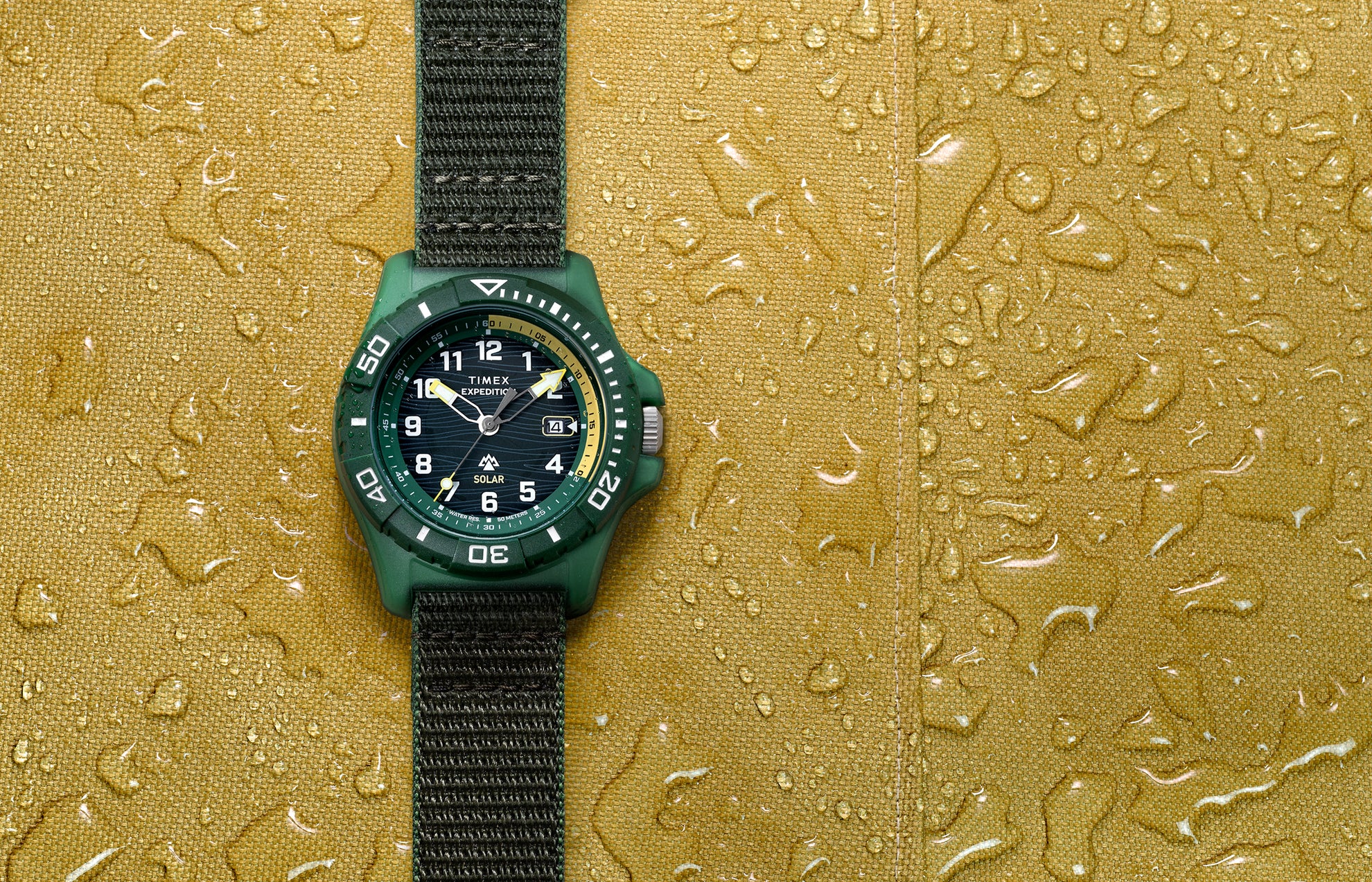 Timex Expedition® Freedive Solar watch with green resin case and black fabric strap displayed on yellow water-resistant surface with water droplets.