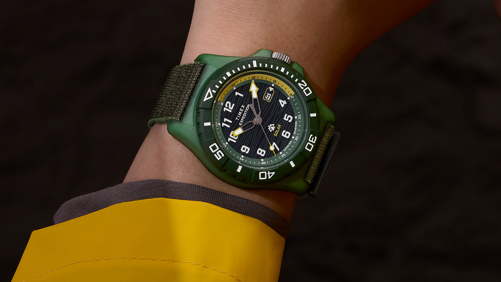 Timex Expedition® Freedive Solar watch with green case and black dial worn on wrist with yellow rain jacket, showing black fabric strap and bold white markers.