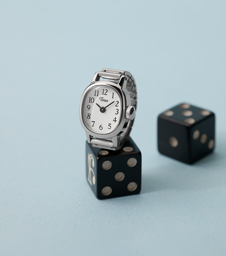 Timex Cavatina® Mini silver-tone watch with white dial displayed on black dice against soft blue background.