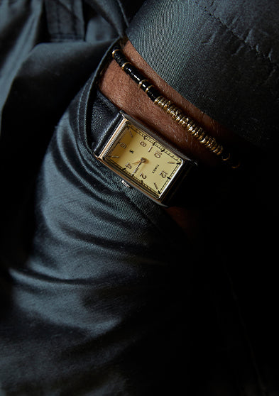 Close-up of a rectangular Timex x Todd Snyder 1976 Lexington watch with a cream dial and black leather strap, worn on a wrist with a dark jacket and beaded bracelet.