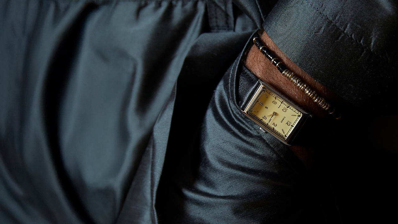 Close-up of a rectangular Timex x Todd Snyder 1976 Lexington watch with a cream dial and black leather strap, worn on a wrist with a dark jacket and beaded bracelet.