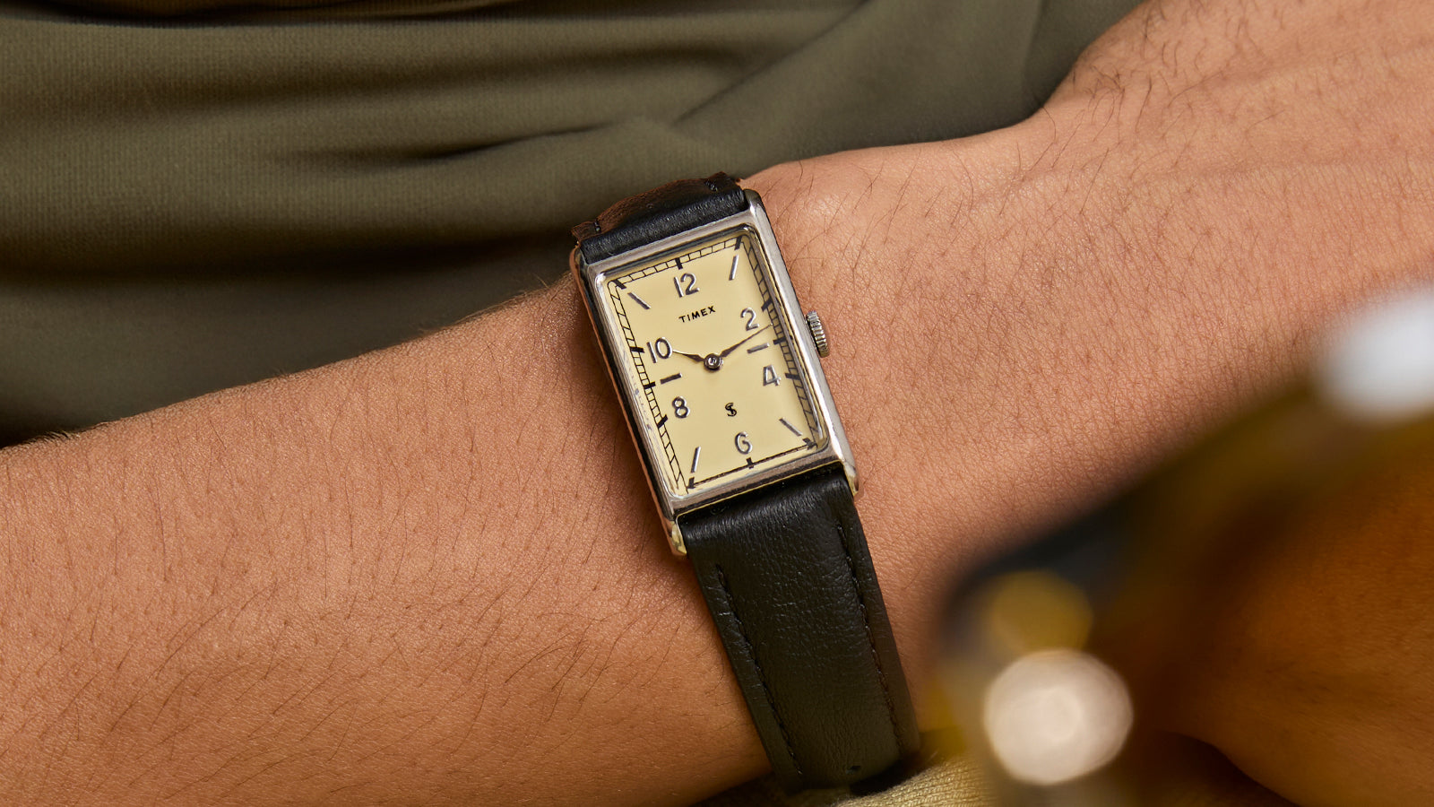 Timex x Todd Snyder 1976 Lexington rectangular watch with cream dial and black leather strap worn on a wrist in a casual setting.