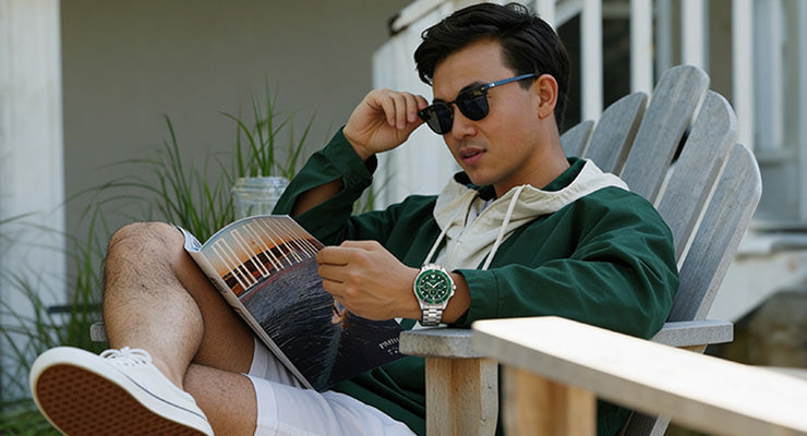 Man wearing sunglasses and a green jacket sitting on an outdoor chair, reading a magazine, with a green watch visible on his wrist.