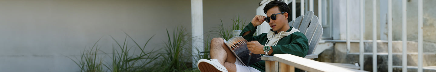 Man wearing sunglasses and a green jacket sitting on an outdoor chair, reading a magazine, with a green watch visible on his wrist.