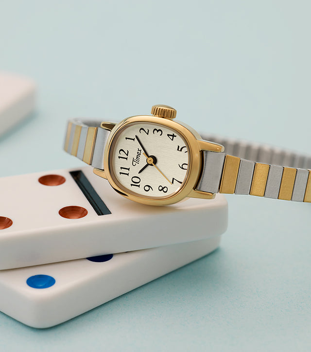 Cavatina® Mini women’s watch with gold-tone case and two-tone stainless steel expansion bracelet resting on stacked dominoes.