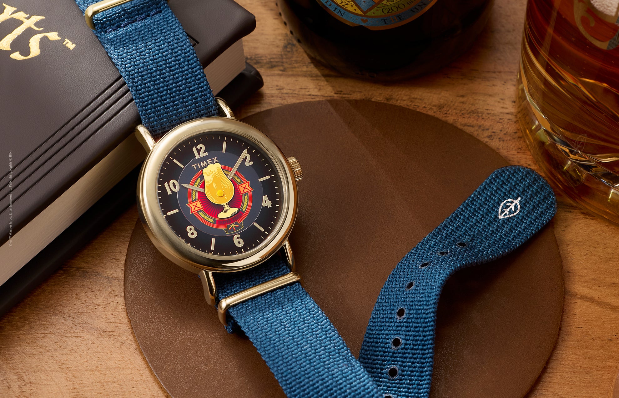 Harry Potter x Timex Weekender Butterbeer watch with gold-tone case and blue fabric strap displayed beside books and a Butterbeer-inspired drink.